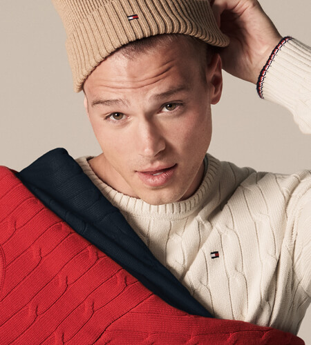 Young model wearing Tommy Hilfiger tan beanie and white sweater while holding matching red and navy sweaters.