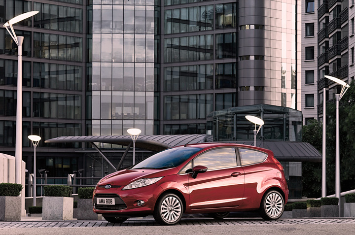 Ford Fiesta vermelho estacionado em frente a prédios comerciais e refletindo luz solar