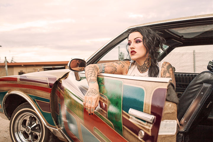 Jovem mulher maquiada e tatuada sentada em carro com pintura customizada com a porta aberta