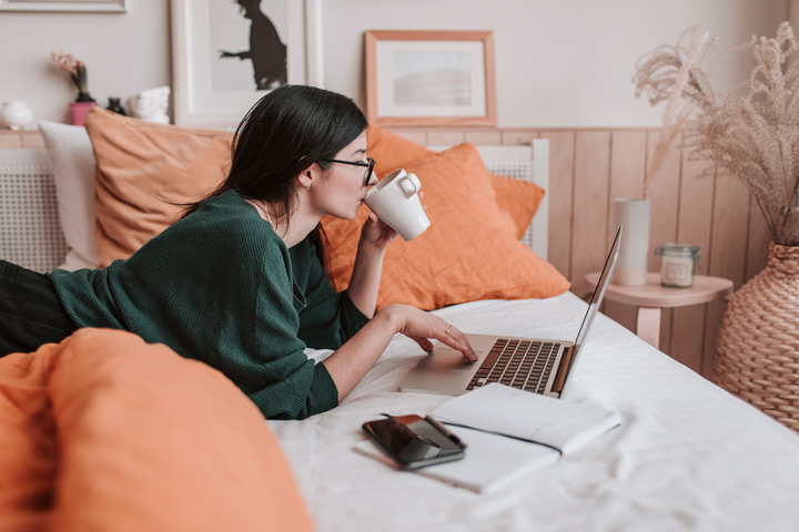 Mulher relaxando na cama bebendo em uma xícara e usando notebook