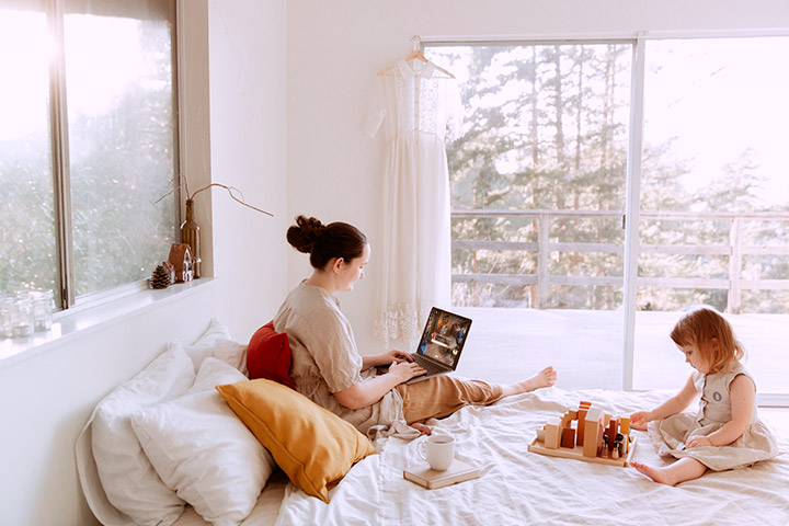 Mãe acessando site da CARUPI em seu laptop sentada em cama de quarto iluminado pela manhã m companhia de filha bebê brincando com bloquinhos