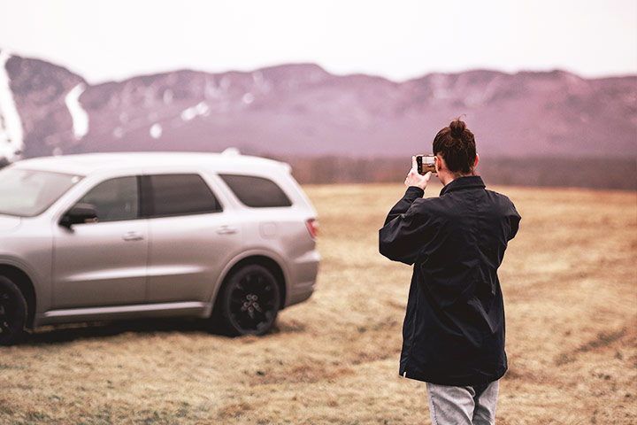 Mulher em ambiente externo fotografando carro com celular em dia nublado com montanhas ao fundo