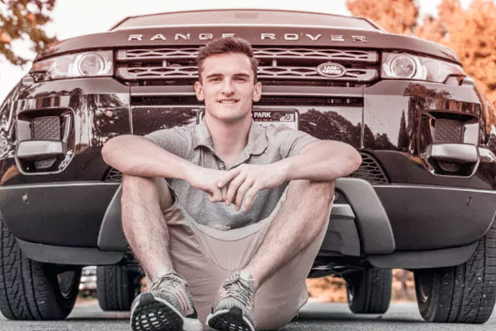 Jovem rapaz sorridente sentado no asfalto em frente a carro Range Rover