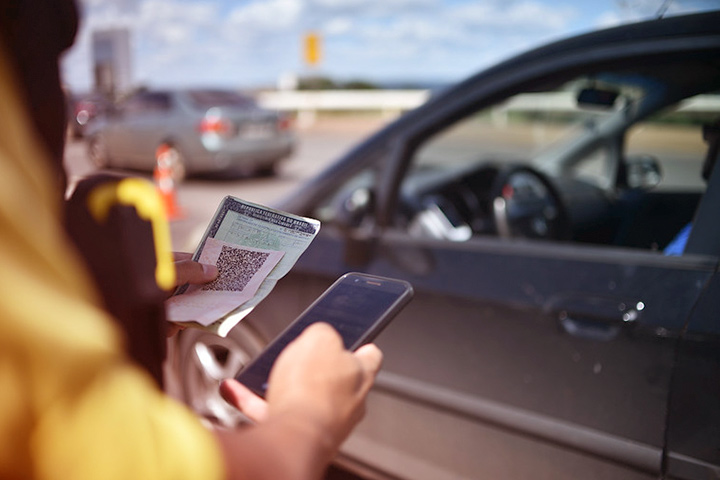 Close em agente de trânsito segurando CNH de motorista em uma mão e checando informações no celular com a outra