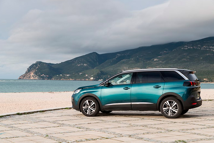 Carro de 7 lugares Peugeot 5008 em tom azulado estacionado em calçada em frente a praia com mar e montanhas ao fundo