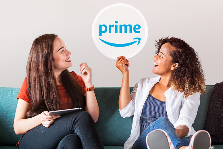 Duas jovens mulheres sentadas em sofá com tablet e segurando uma placa com logomarca da Amazon Prime