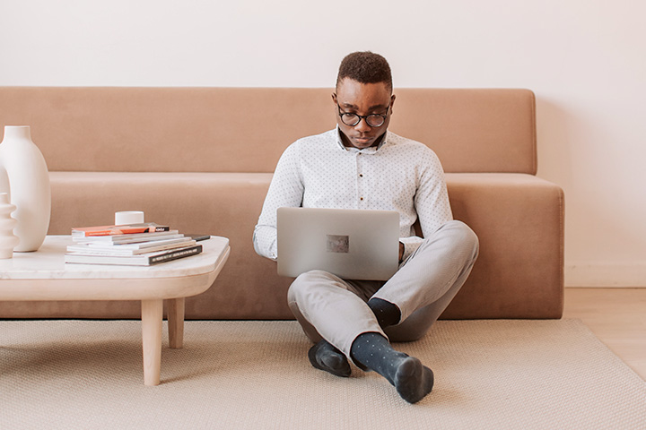 Jovem afro-americano sentado no tapete da sala de TV acessando website CARUPI em seu laptop