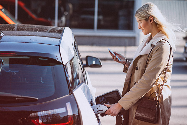 Jovem mulher carregando seu carro elétrico enquanto observa a tela de seu celular