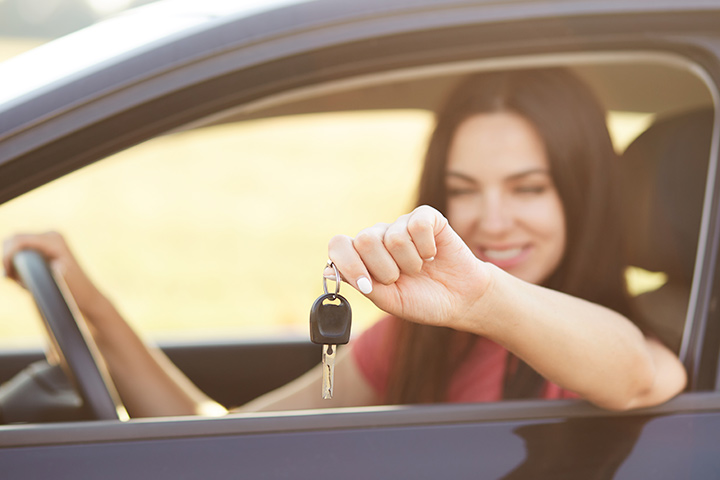 Jovem mulher sorrindo sentada em carro com mão ao volante e braço para fora da janela mostrando chave do veículo