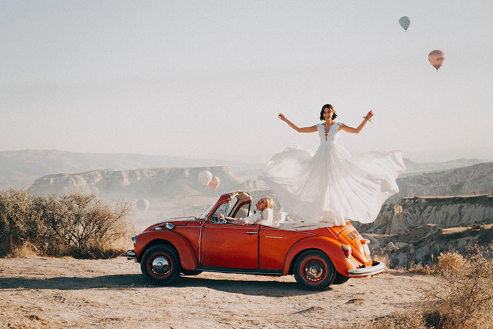 Fusca vermelho conversível parado à beira de um desfiladeiro em cenário desértico com noiva em pé no banco de trás e vestido esvoaçante