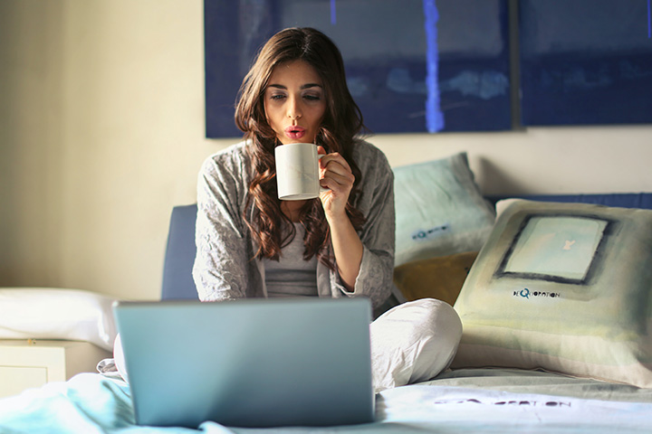 Moça relaxando em sua cama bebendo café em xícara e acessando site da Carupi em seu laptop