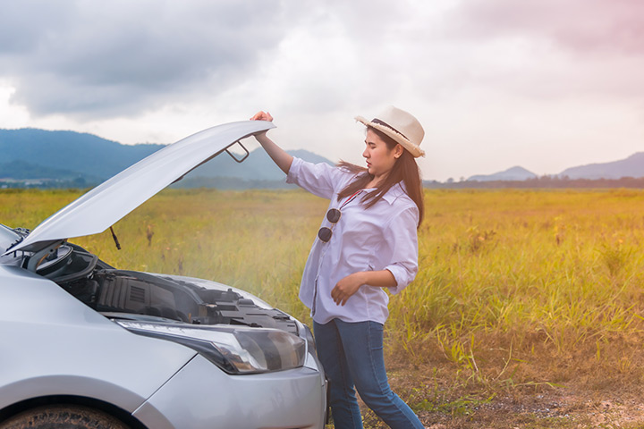 Jovem mulher abrindo capô de seu carro com motor superaquecido em acostamento de estrada com paisagem natural ao fundo