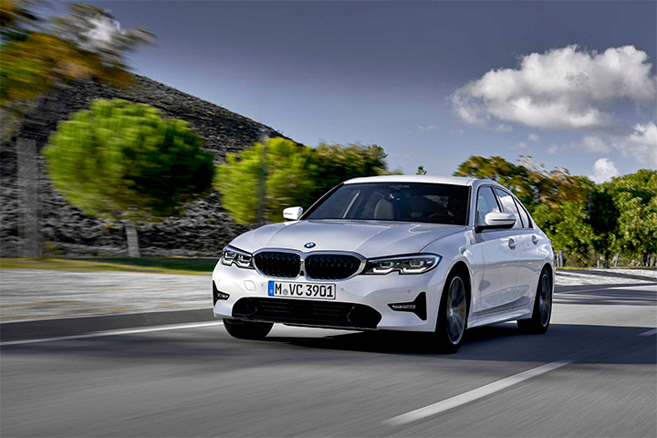 BMW 320i na cor branca trafegando em via urbana arborizada com céu azul ao fundo