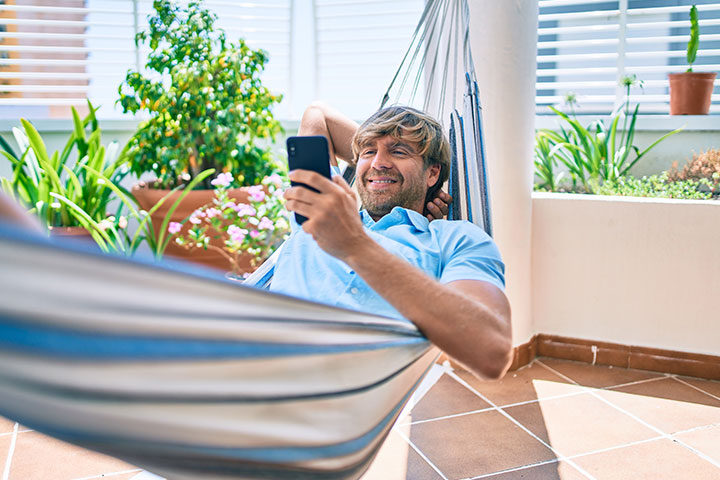 Homem sorrindo e relaxando em rede pendurada na varanda e acessando site da Carupi em seu celular