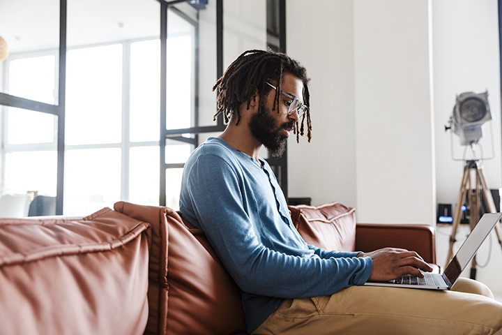 Jovem homem afro-descendente sentado em sofá de casa acessando site da Carupi em laptop em seu colo