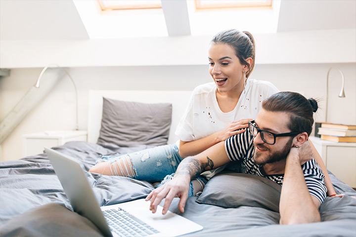 Jovem casal com visual moderno deitado de forma descontraída na cama acessando site da Carupi em notebook