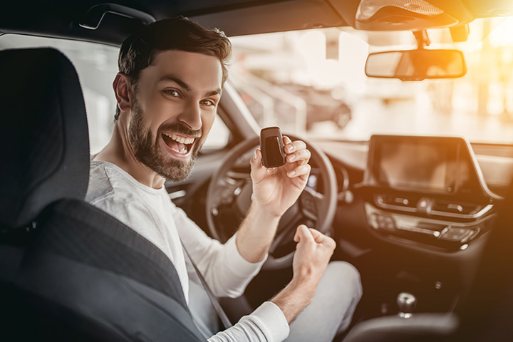 Jovem rapaz feliz dentro de carro mostrando a chave do veículo e comemorando a aquisição