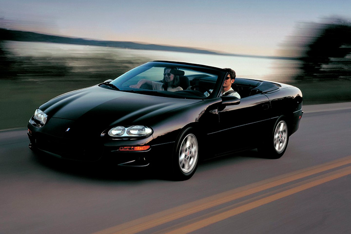 Chevrolet Camaro conversível preto ano 2001 com casal dirigindo em alta velocidade em estrada com fundo desfocado