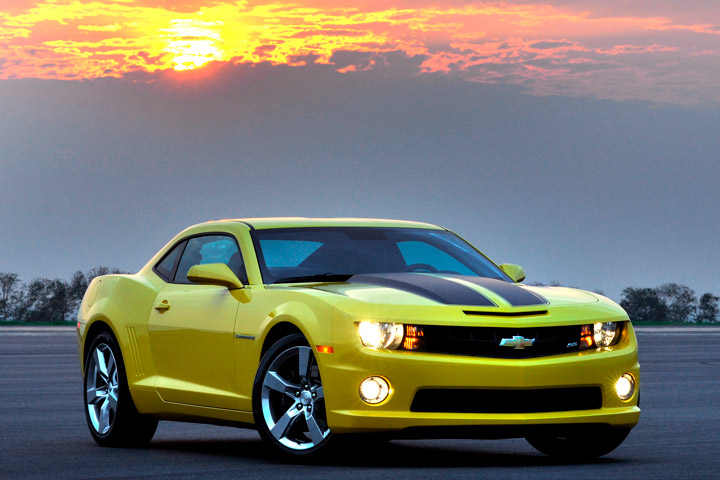 Chevrolet Camaro amarelos com faróis acesos em pátio exterior com sol se pondo atrás das nuvens