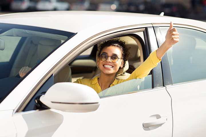 Jovem mulher dirigindo carro feliz e fazendo gesto de positivo com braço para fora da janela