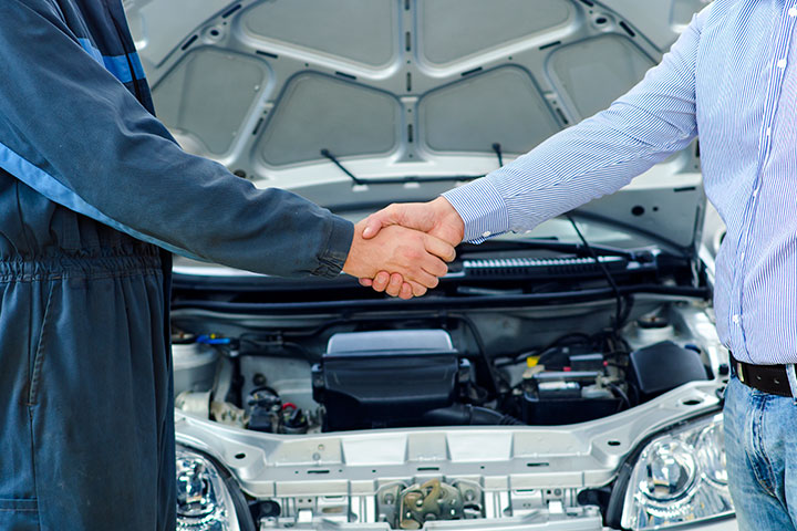 Mecânico e cliente dando um aperto de mão com carro ao fundo com capô aberto