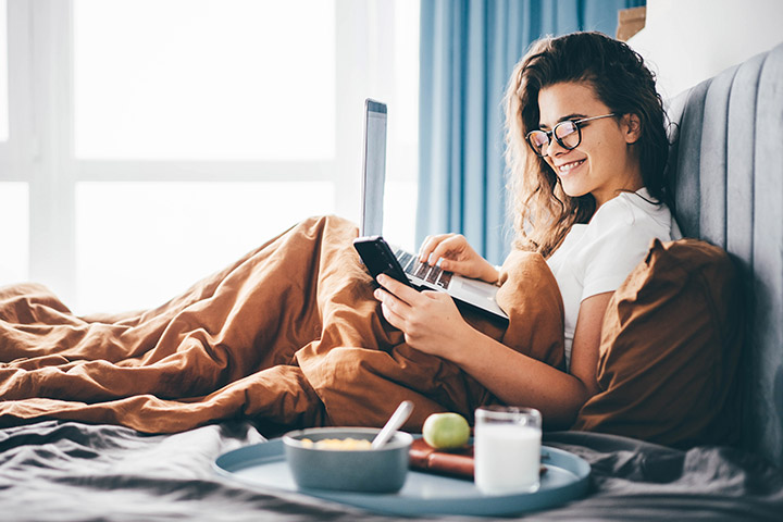 Jovem mulher tomando café da manhã em sua cama e acessando plataforma da Carupi em celular e notebook