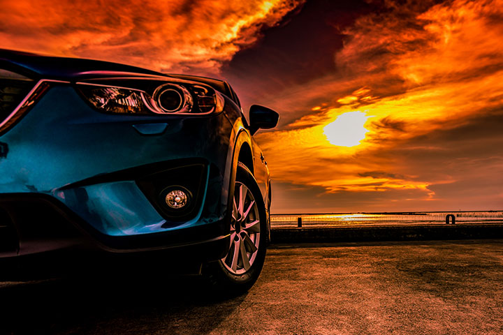 Carro azul estacionado em frente ao mar refletindo cor amarela intensa do sol no horizonte