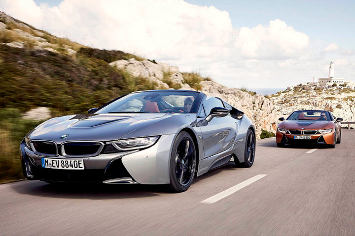 Dois carros BMW i8, um prateado e outro alaranjado, em rodovia costeira com paredão rochoso ao lado