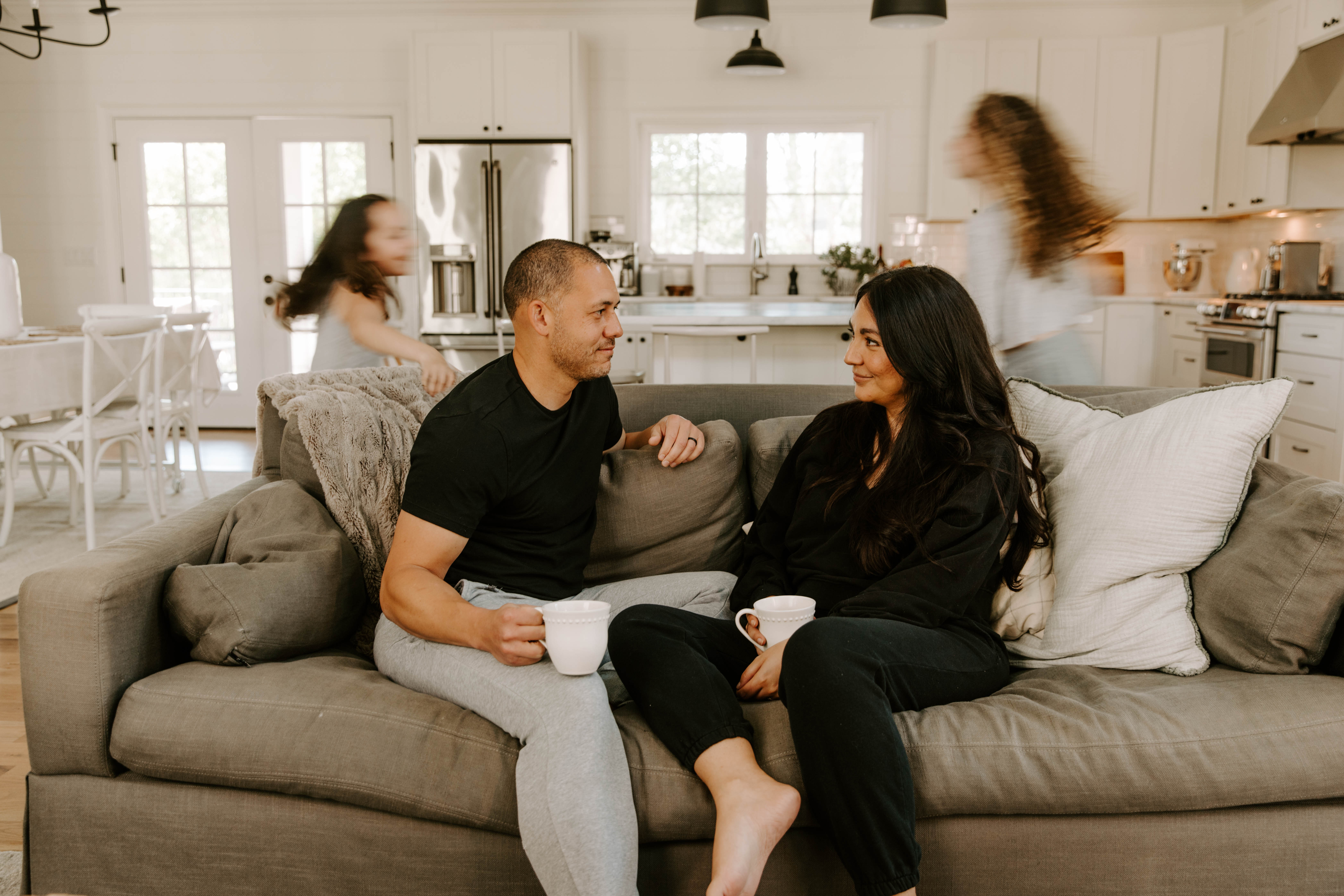 A couple relax on the sofa while their kids run around, playing, behind them.