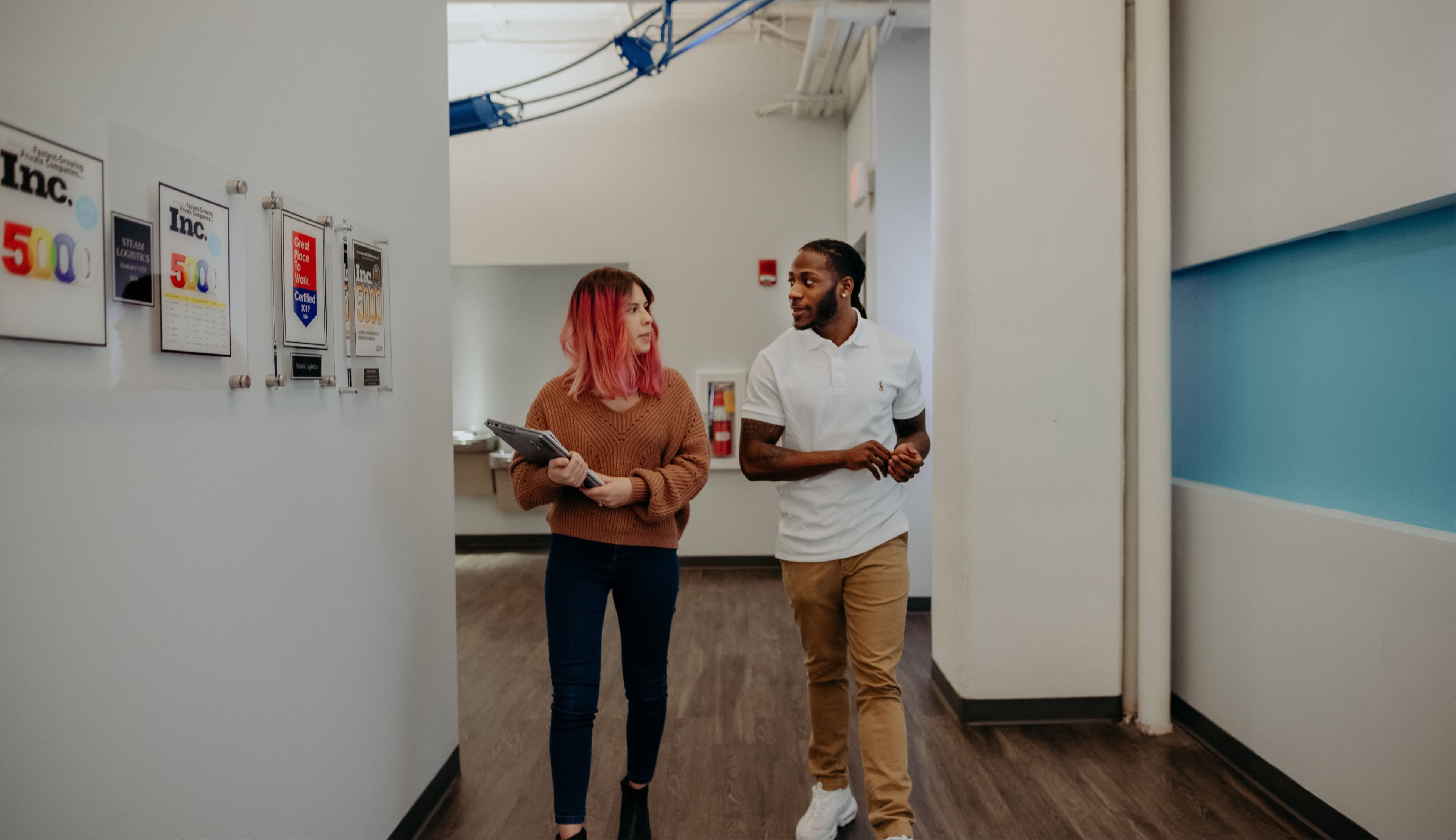 Two Steam employees walk down a hallway with some of the company's accolades, like Inc. 500, framed on the wall.