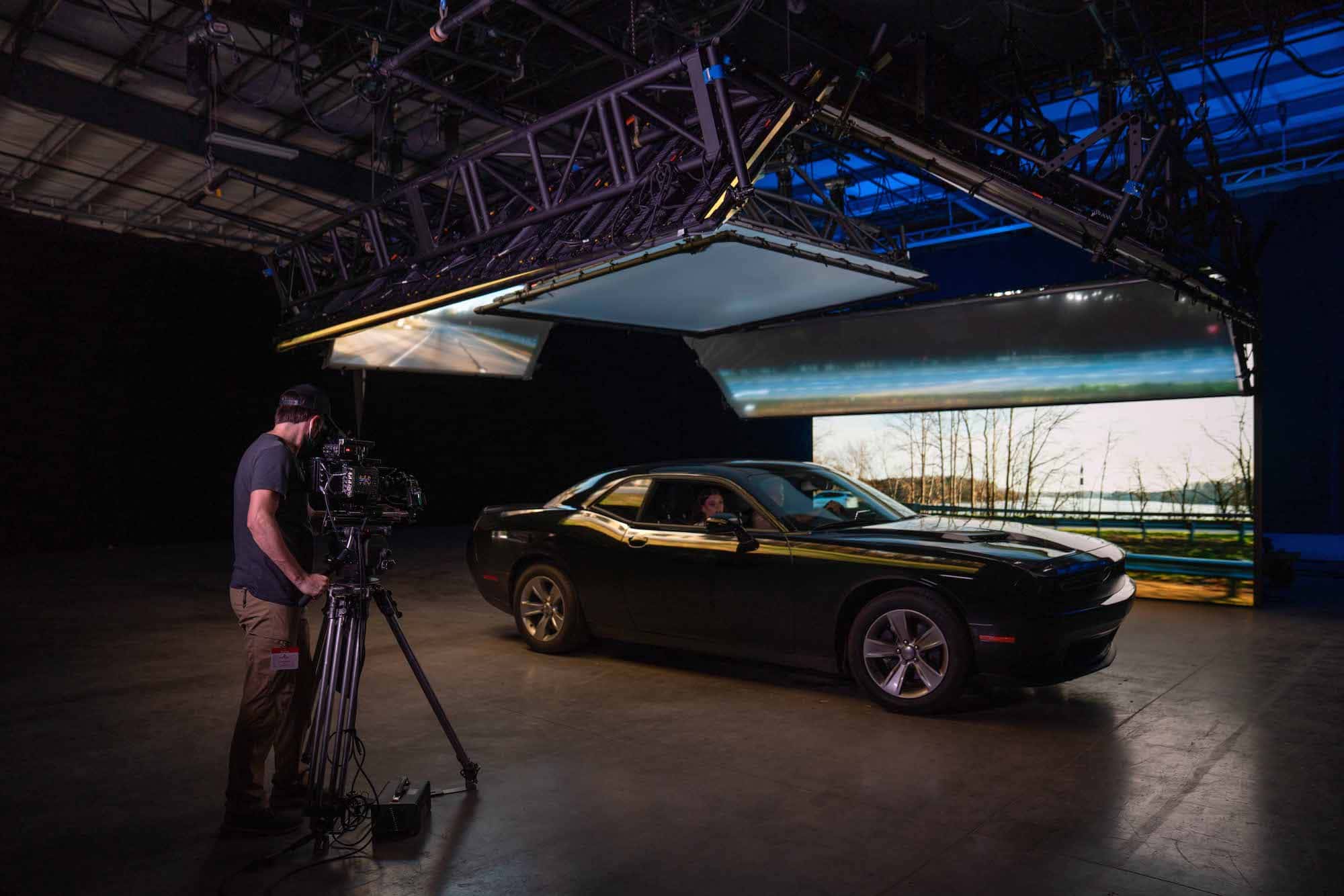 Medium shot of a man filming a car inside of a studio stage. A screen behind the car displays a landscape, mimicking the movement of the car.