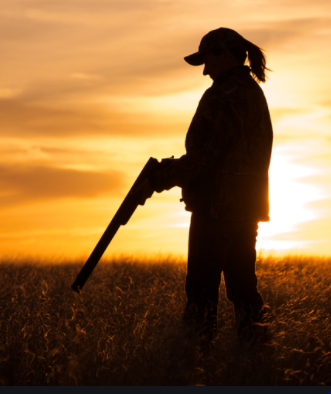 Woman with shotgun in front of sunset.