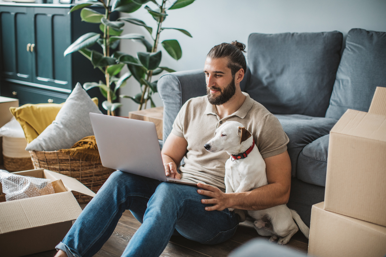 man sitting on floor by dog after unpacking checking his laptop for customer reviews for his sales training business | Profi