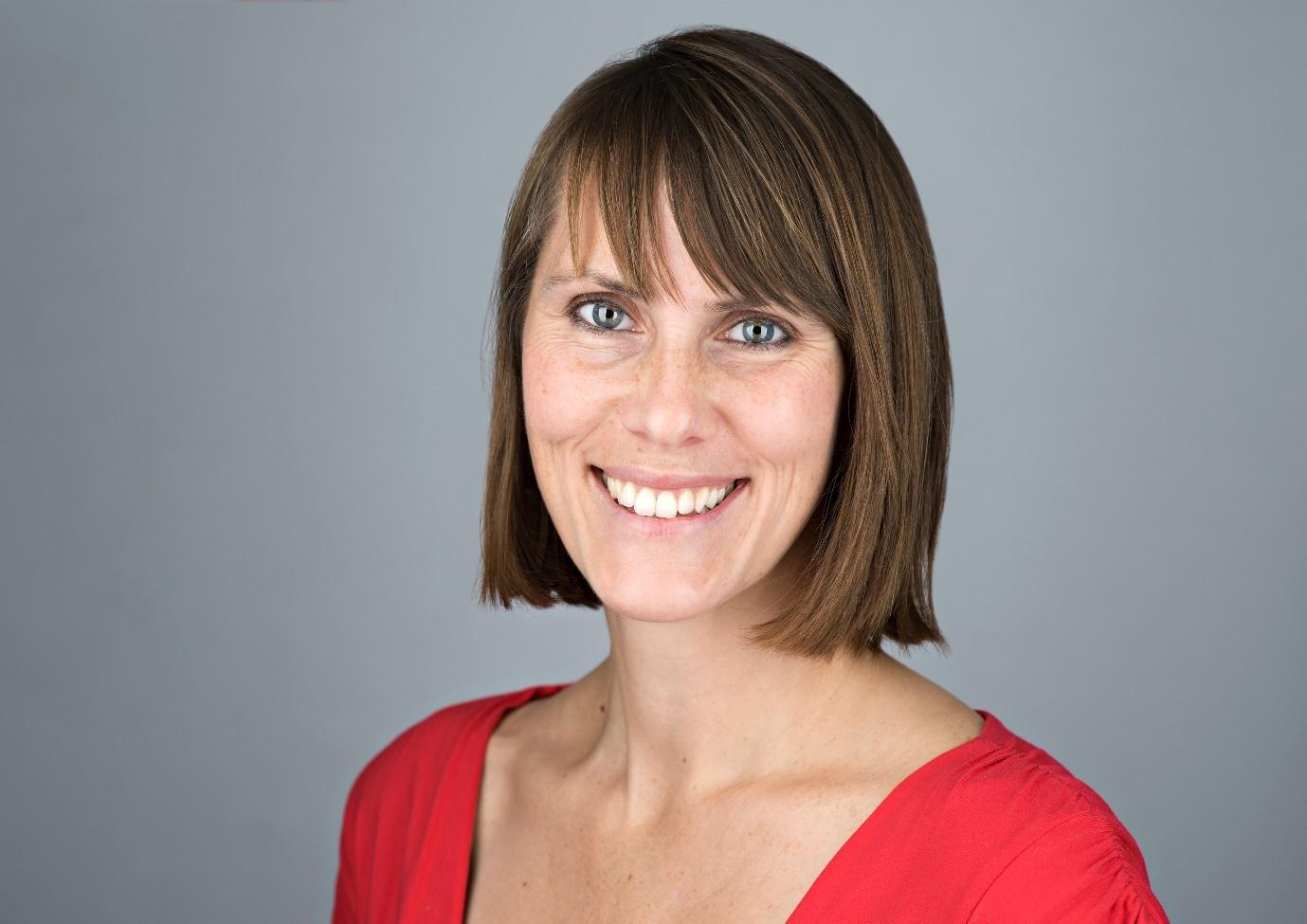 Suzy smiling into camera in front of a grey background. She is wearing a red top.