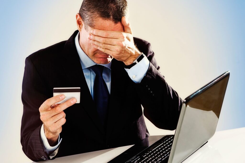 A man in a suit at his laptop with credit card in-hand; holding his other hand over his face to represent trust in his purchase