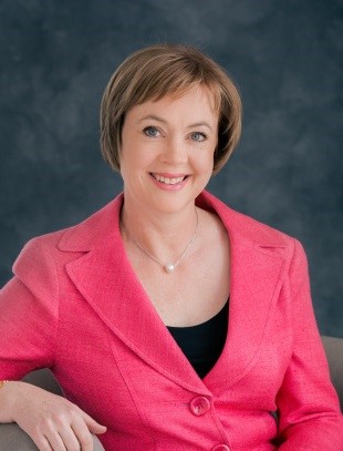 Woman smiling and sitting on a chair. She is wearing a pink suit.