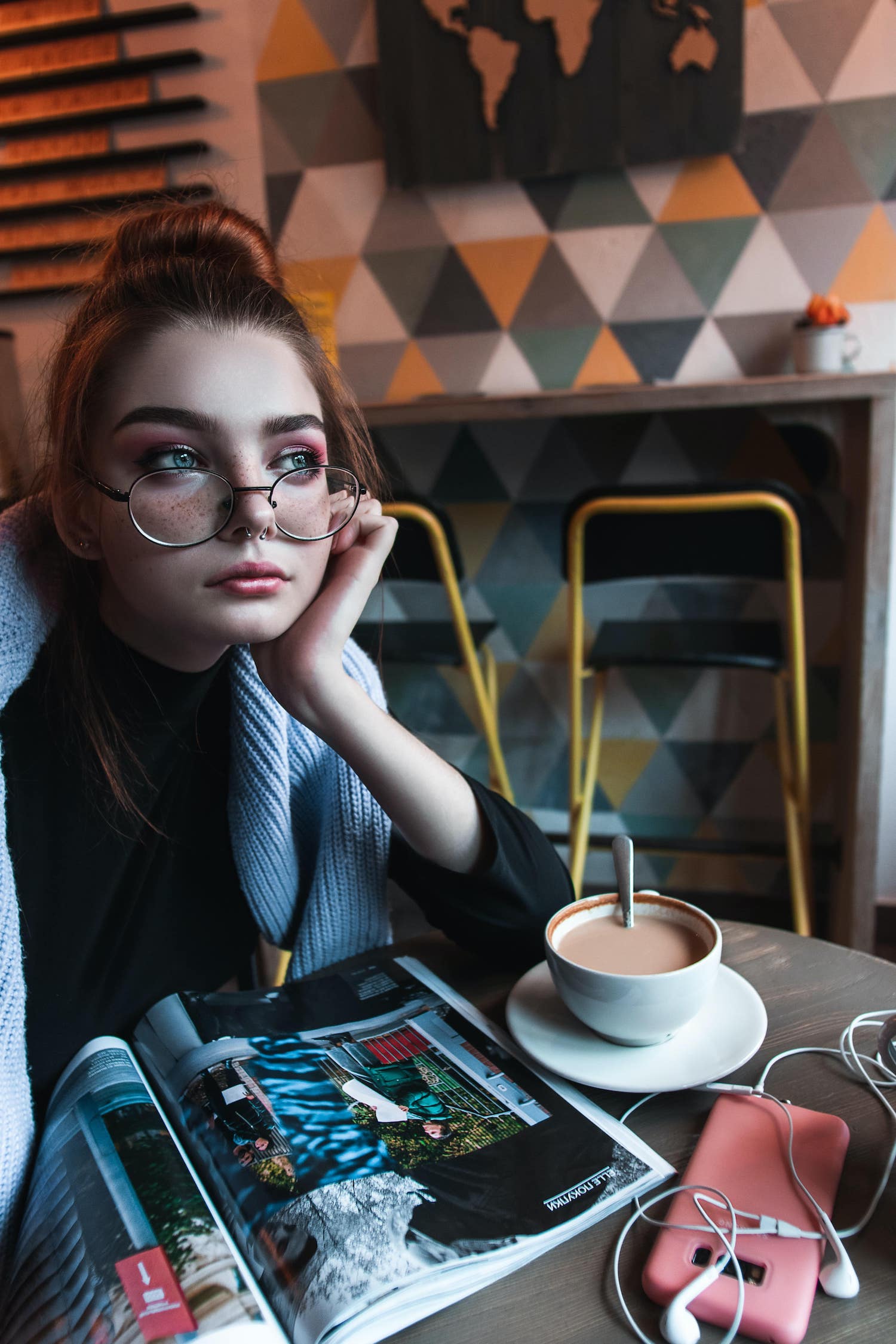 A young girl wearing glasses in a café, drinking a coffee and reading a magazine.