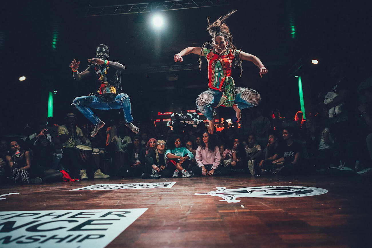 Two street dancers jumping in a dance battle surrounded by a crowd.