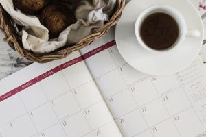 Full cup of tea sitting on an open calendar, nect to a wicker basket full of bread rolls.