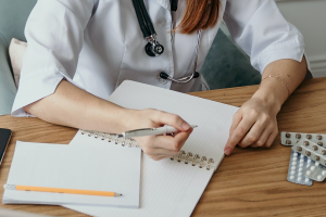 Doctor with a pen in hand, about to write in a notebook, with pills next to their left arm.