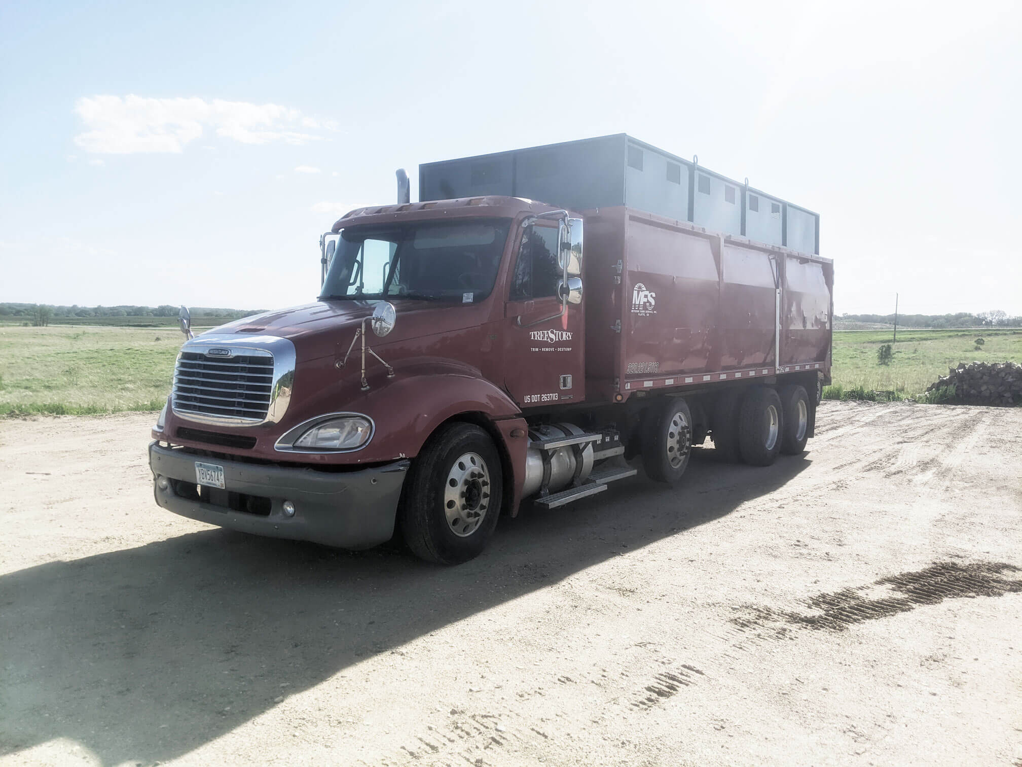 Red Freightliner Chip Trucks parked on gravel lot with vegetation behind. This equipment lets TreeStory leave jobsites clean