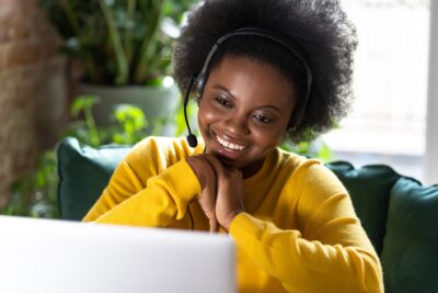 A woman in a headset smiles at her laptop as she watches a virtual panel discussion of nonprofit coaching consultants..