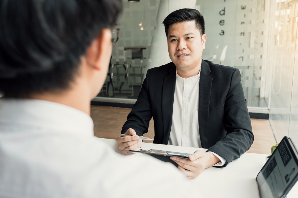 Two men face each other an interview for a nonprofit organization. One faces the camera, asking a question. We see the silhouette of the other's head and shoulder in the left foreground.