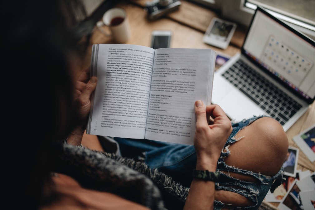 This photo was taken over a woman's shoulder. She holds a book open, reading, as she sits cross-legged in ripped jeans in front of her laptop.
