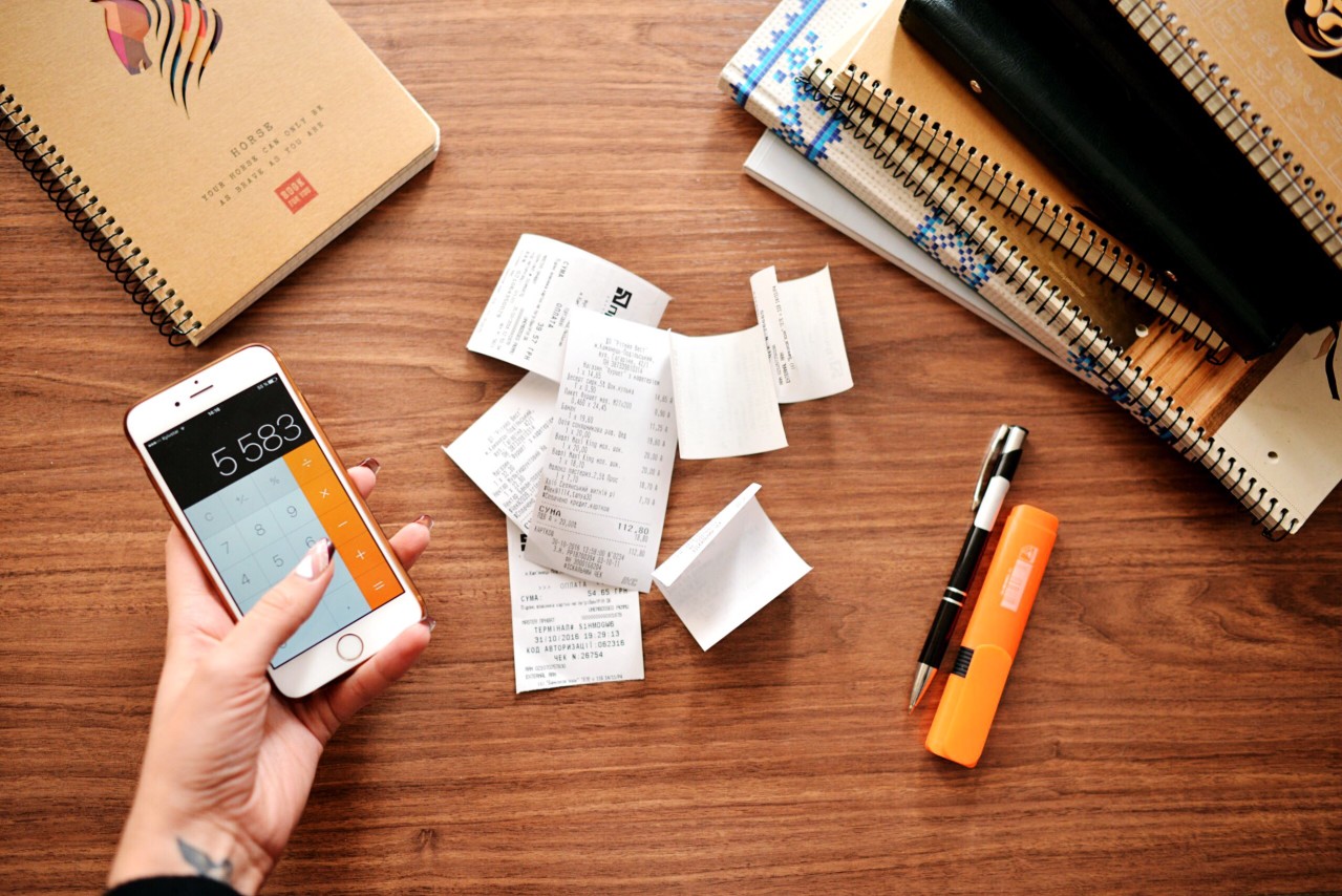 Fiscal sponsorship is a good option for new nonprofits. In this photo, we see a flat lay of a hand holding a cell phone with the calculator app open; a pile of receipts, and various writing utensils against a wooden desk.