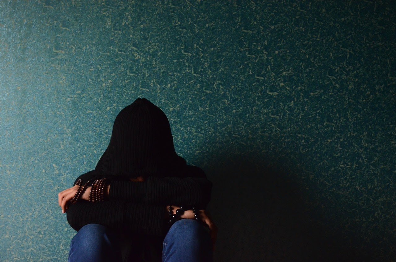 A woman sits against a wall with her face in her arms, appearing to be depressed or in some sort of emotional distress.