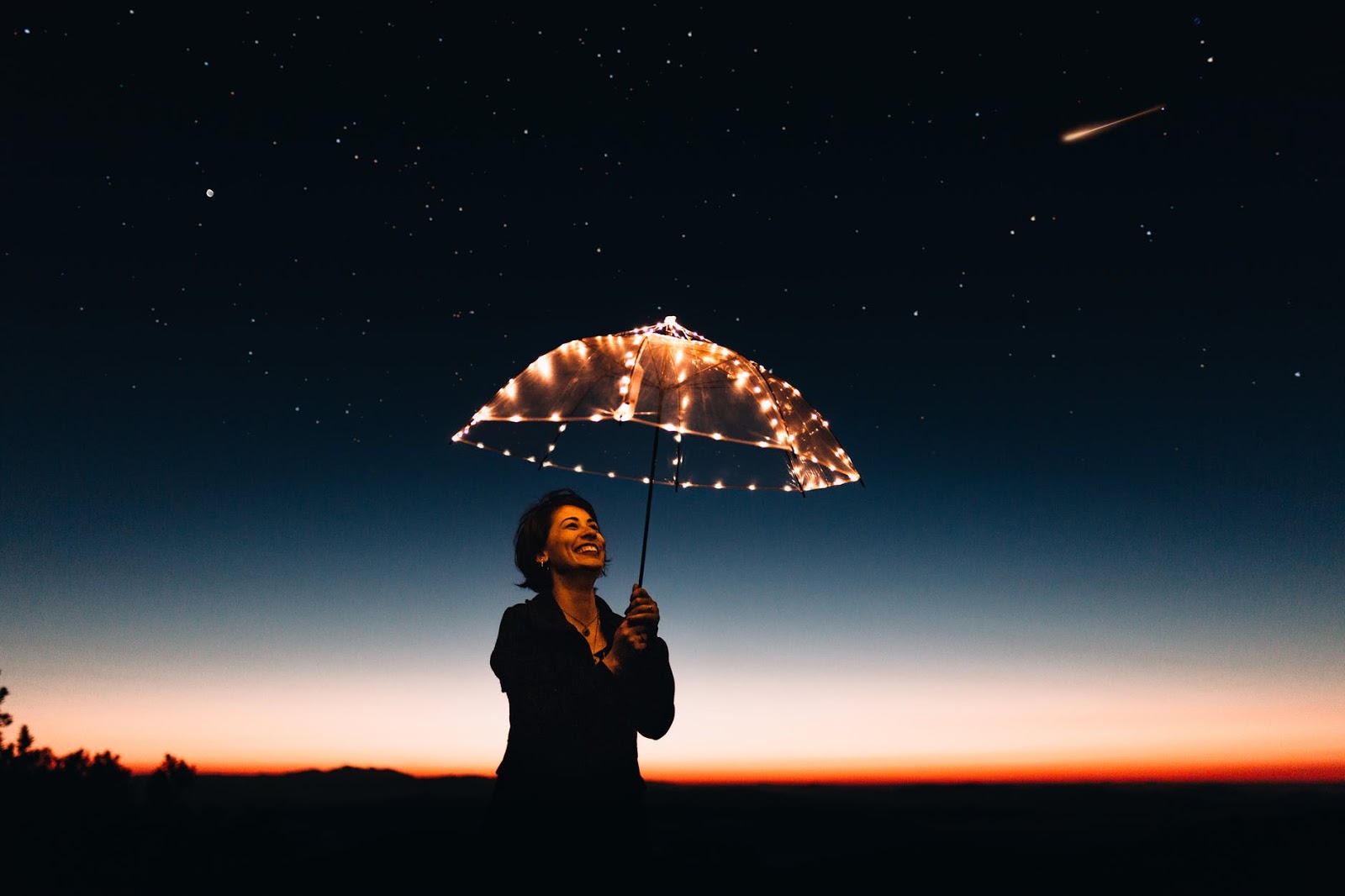 A woman, smiling, holding an illuminated umbrella at dusk