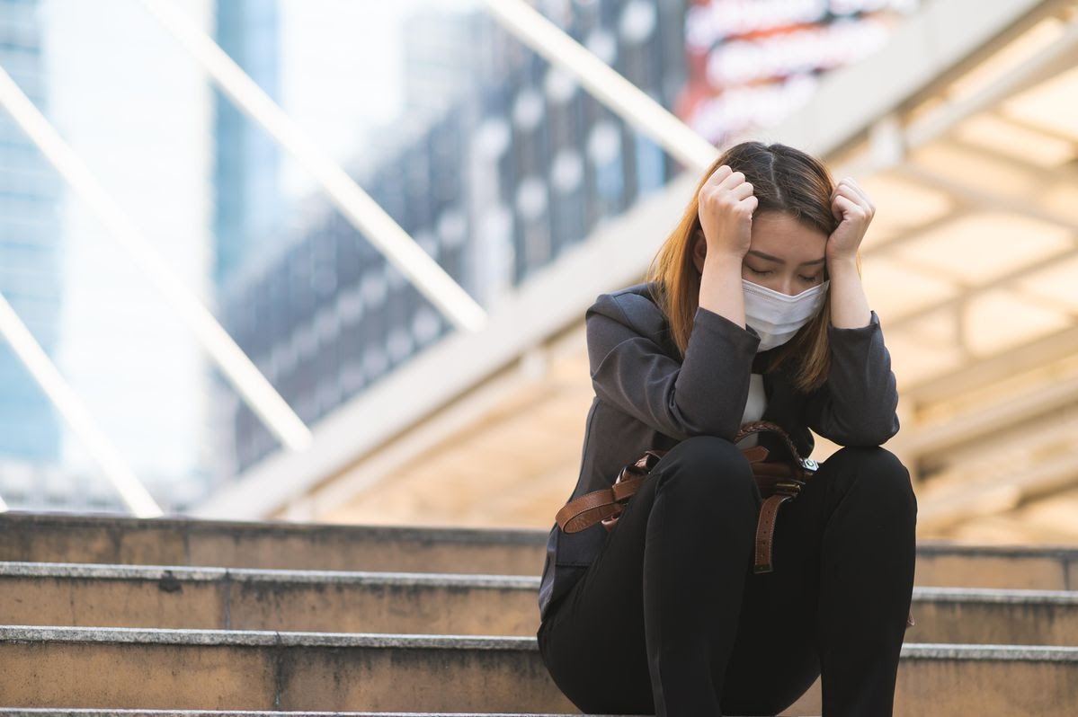 A woman wearing a mask sits outdoors on concrete steps while holding her head in her hands.