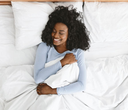 An overhead photo of a happy-looking woman, snuggled in a comfortable bed with crisp white linens