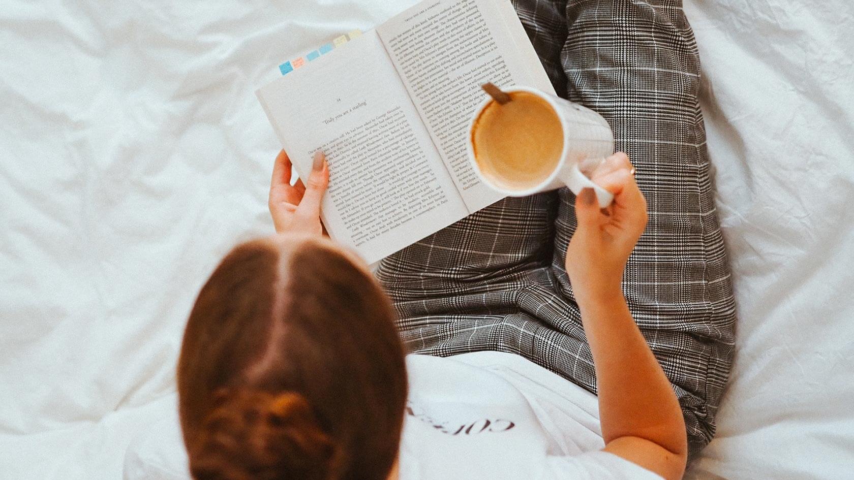 A woman sitting on her bed reading a book with a cup of coffee